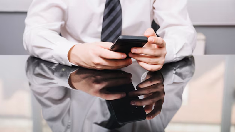A Businessperson in a white shirt and striped tie using a smartphone at a reflective glass table.
