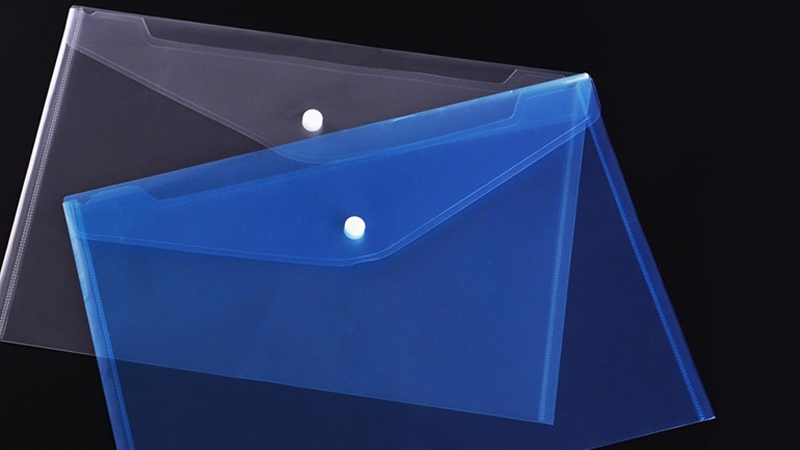 A clear colorless envelope layered over a translucent blue one, both placed on a black background, with a pile of colorful jotters partially visible at the edge of the image.