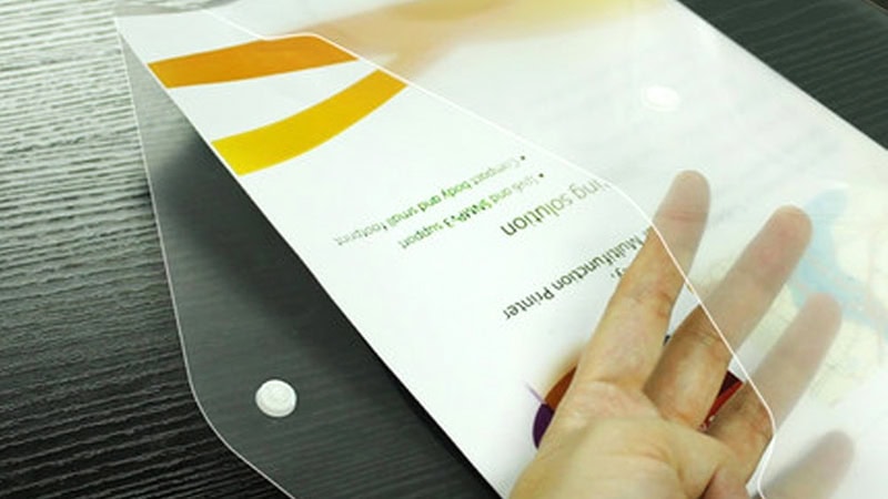 A close-up of a hand opening a clear plastic envelope containing a printed document. The envelope has a snap button closure and is placed on a dark wooden surface.