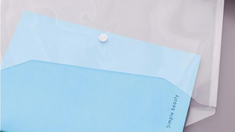 A person is placing a blue-colored document into a clear plastic envelope. A pen and another small object, possibly a jotter, are also visible in the scene, suggesting a workspace setup.