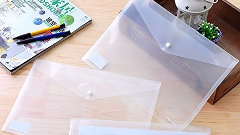 Three clear plastic envelopes are placed on a table. one holds a document while the other two are empty. Nearby, a magazine with two pens rests on the surface, alongside a wooden tray holding a small flowerpot and a white lantern
