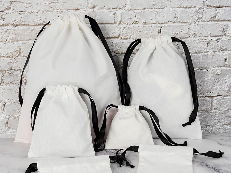 A high-angle shot captures two drawstring pouches on a white marble surface with subtle gray veining. On the left is a larger light blue pouch with light-colored drawstrings. To its right and slightly in front is a smaller, cream-colored pouch with black drawstrings. Both pouches are cinched at the top, and their fabric appears slightly rumpled. Natural light casts soft shadows.