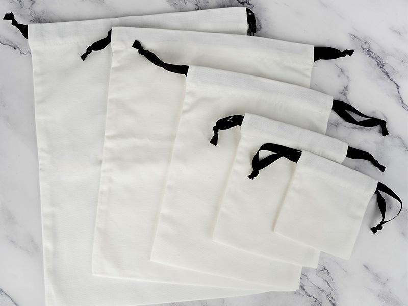 A close-up shot focuses on a cream-colored drawstring pouch adorned with intricate embroidery. The embroidery features delicate light pink roses with green stems and leaves, scattered across the lower portion of the pouch. The top of the pouch is cinched, creating ruffles, and a white drawstring with a single white pearl bead hangs down the left side. The background is softly blurred, keeping the focus on the pouch's details.