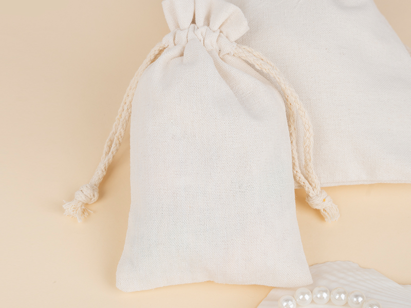 A dimly lit, eye-level shot features two small, natural-colored fabric pouches sitting on a dark wooden surface, possibly a desk or mantelpiece. The pouch on the left is lighter in color and has a small tag hanging from its drawstring that reads "ARCTIUM" vertically. The pouch on the right is darker, a brownish-beige. In the background, a dark, wooden, possibly antique, object or furniture piece with metallic accents is visible. Reflections from a window or light source can be seen in the background on the left.